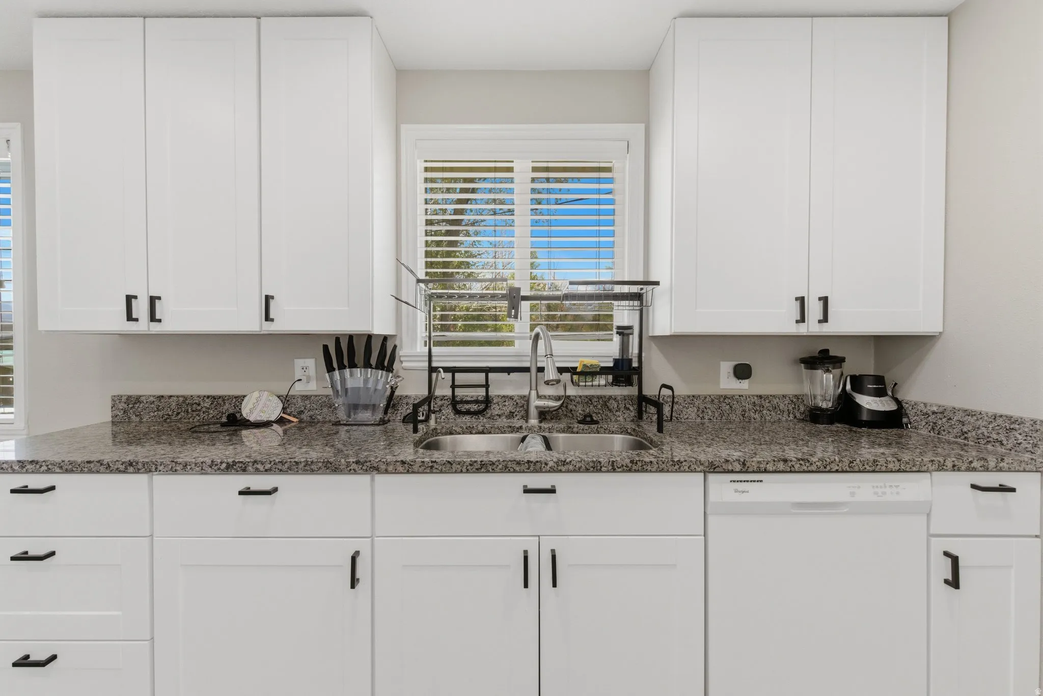 Contemporary kitchen featuring white shaker cabinetry with matte black hardware, speckled countertops, a double basin stainless steel sink, and a white built-in dishwasher