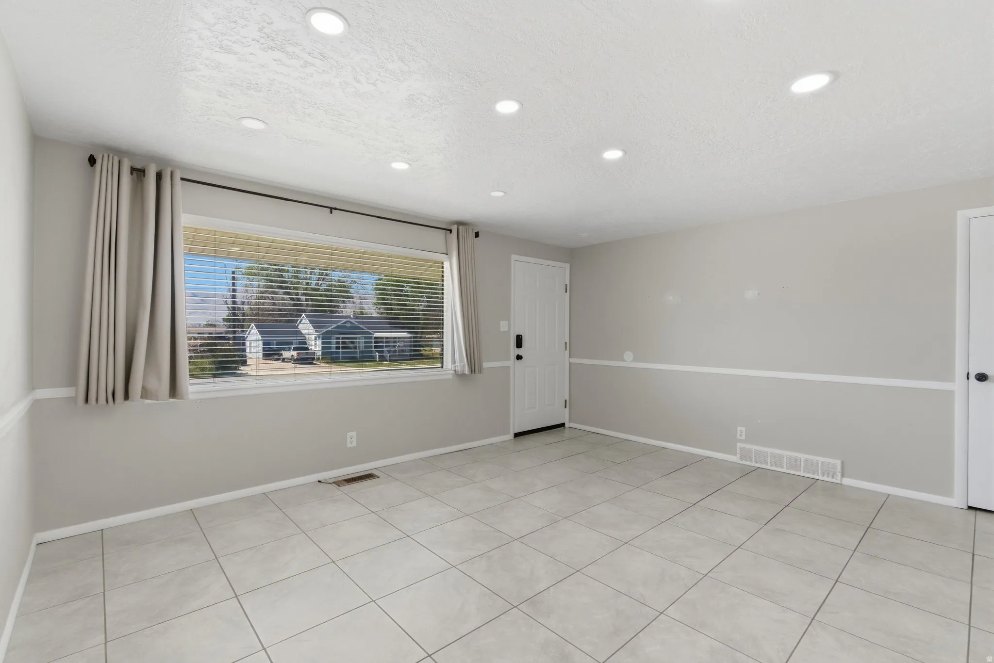 Spacious living area featuring large format tile flooring, recessed ceiling lighting, a prominent front entry door with black hardware, and a wide window with blinds and drapes