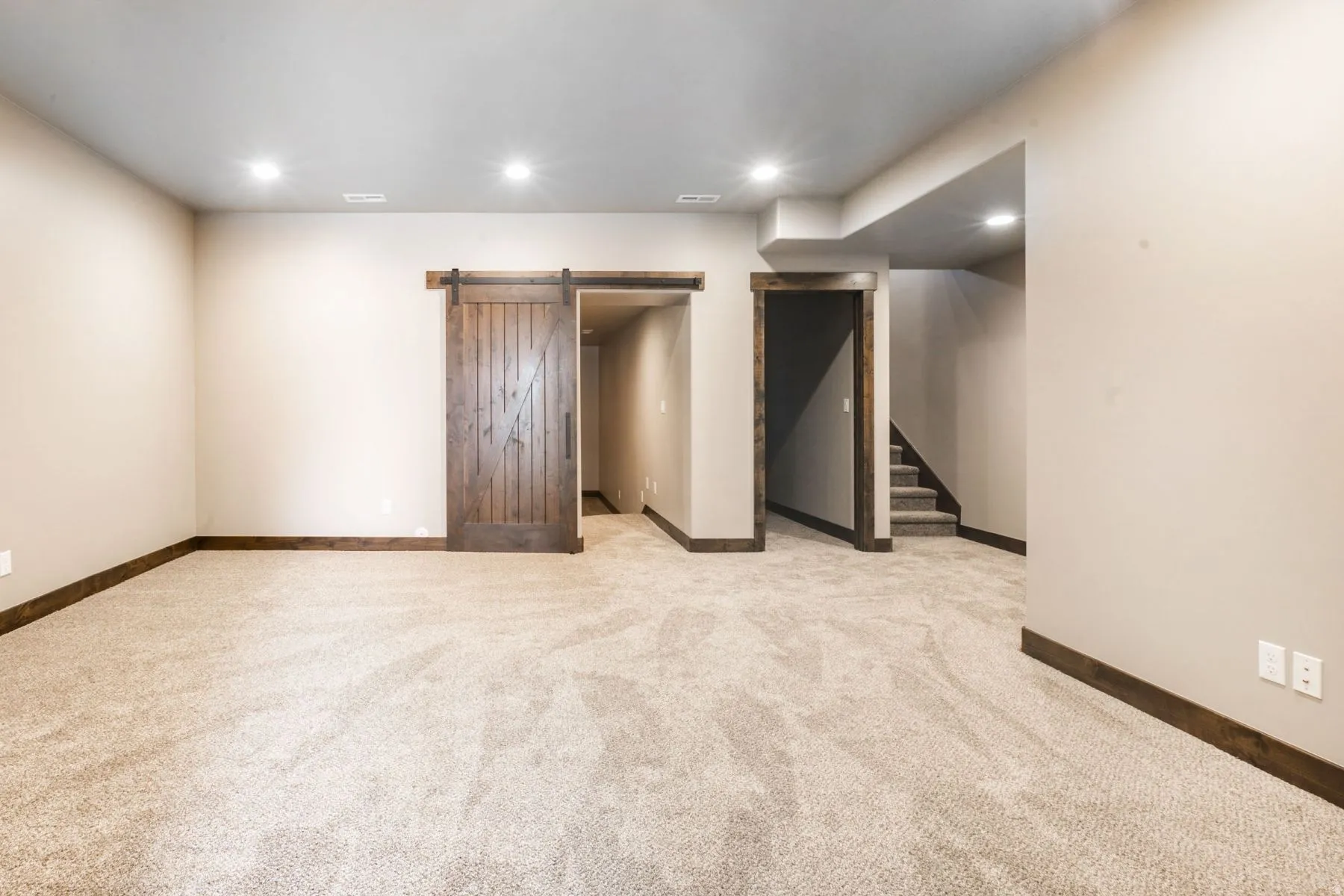 Spacious room featuring neutral carpeting, recessed lighting, a stained wood barn door, wood trim accents, and an open doorway to a carpeted staircase
