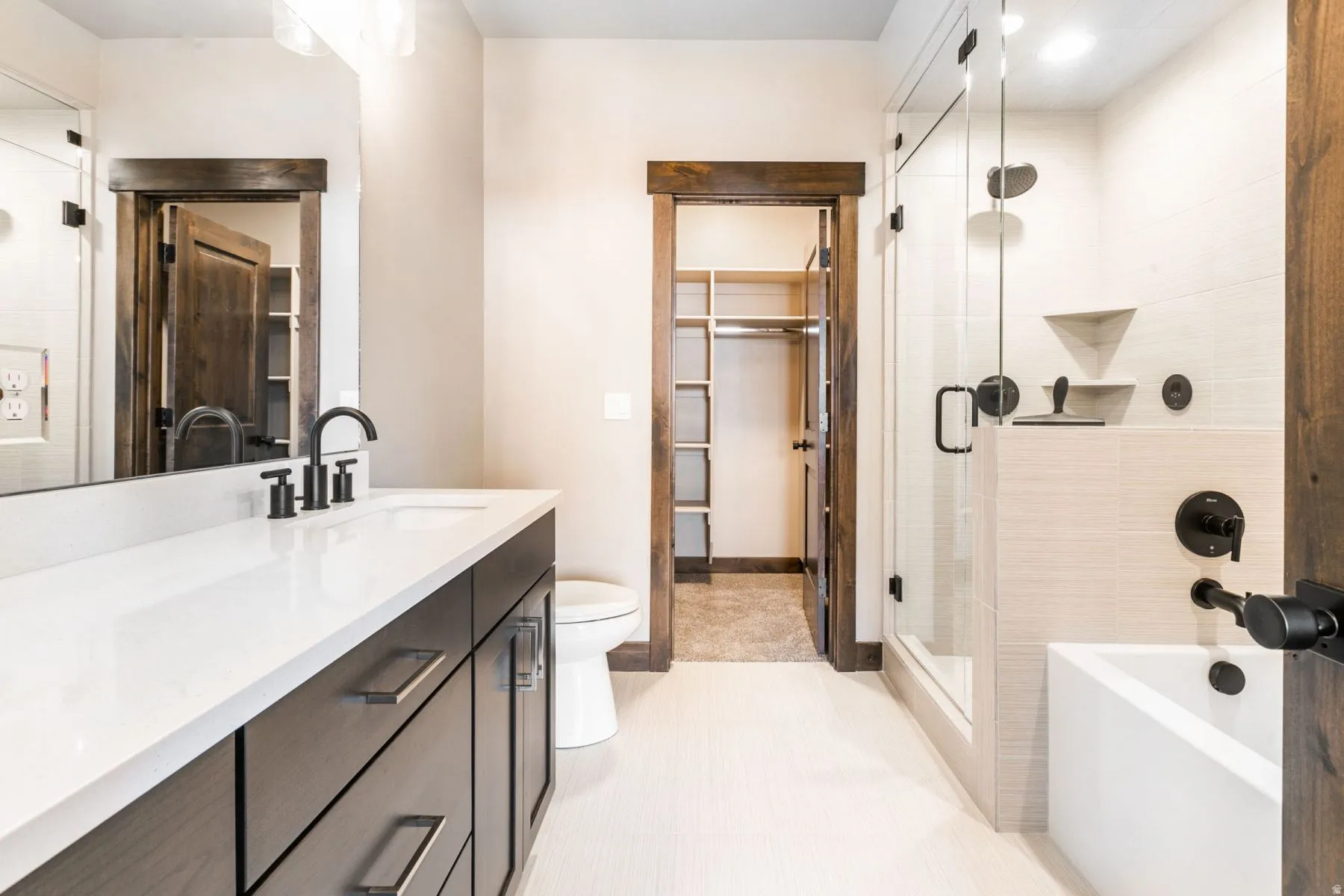 Bathroom featuring a wood-finish vanity with a white countertop and integrated sink, matte black fixtures, a built-in tub with a glass shower enclosure, and a walk-in closet with built-in shelving