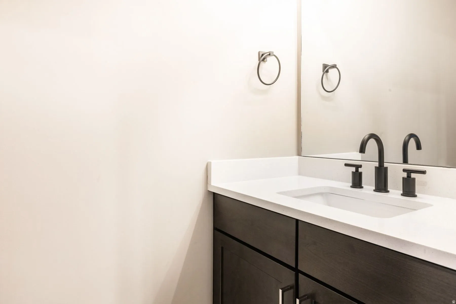 Bathroom vanity featuring a light-toned countertop with an integrated rectangular sink, matte black widespread faucet, and dark wood-finish cabinetry