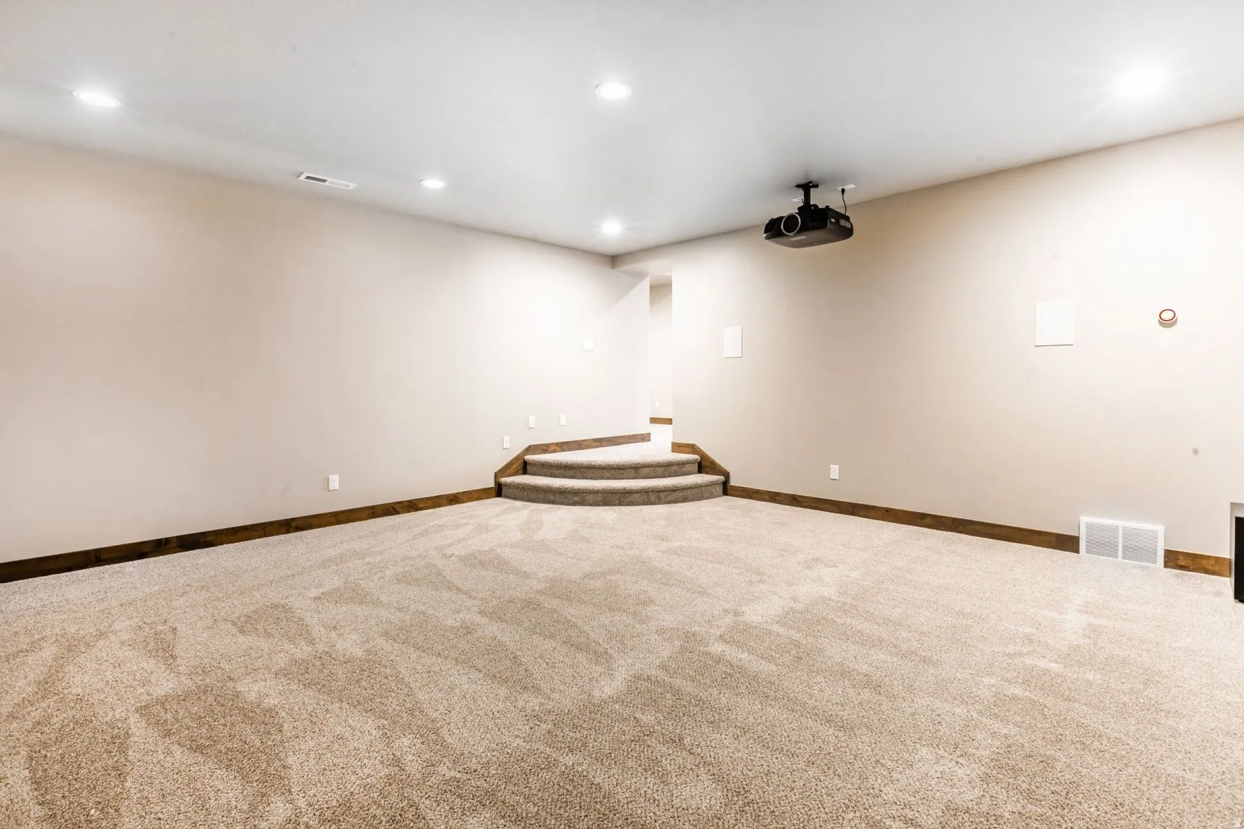 Spacious room featuring neutral carpeting, a ceiling-mounted projector, recessed lighting, and a built-in tiered carpeted platform