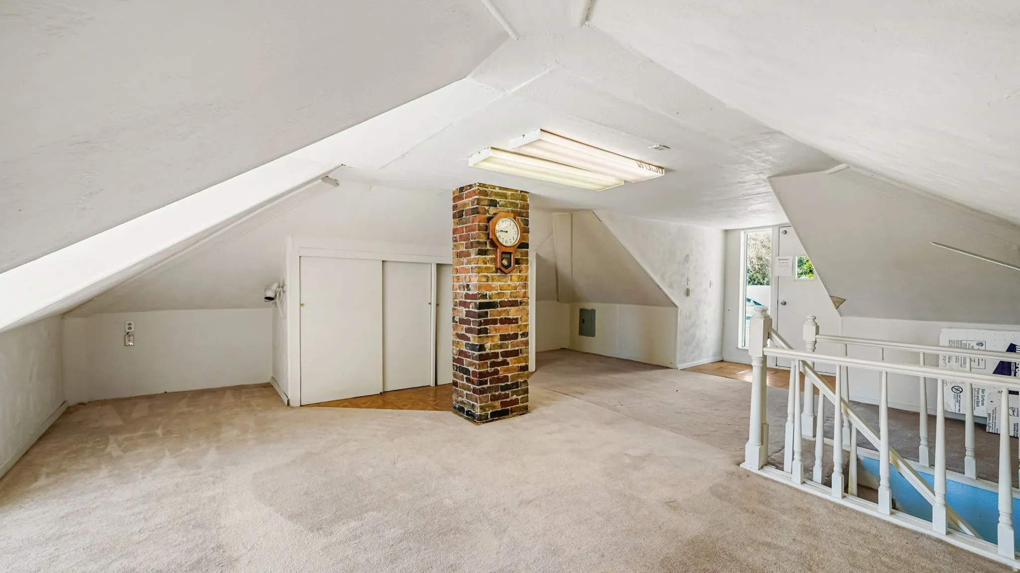 Expansive attic space featuring sloped ceilings, a brick column, recessed storage with sliding doors, a built-in stair railing, and an overhead fluorescent light fixture