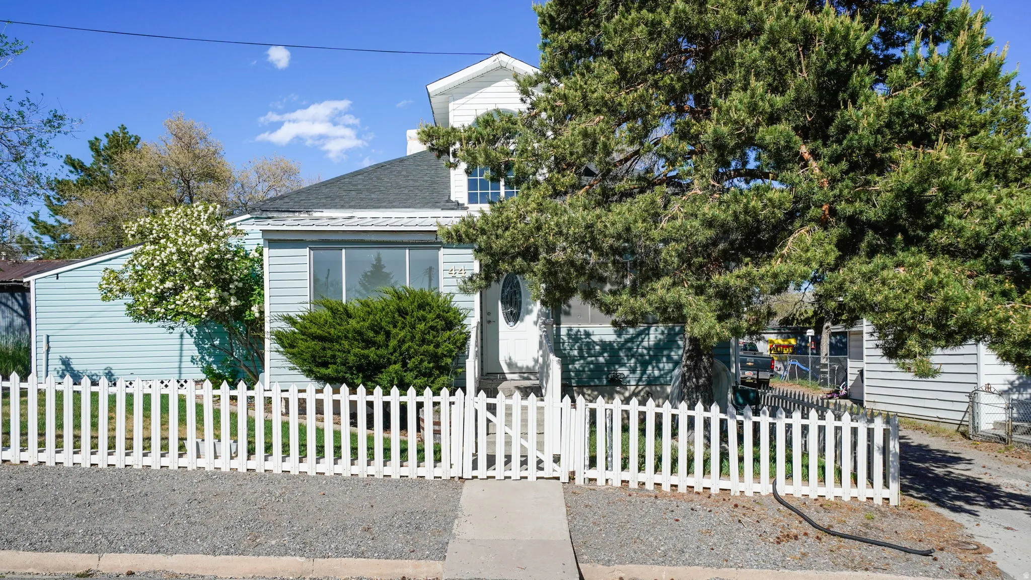Light blue siding exterior with white picket fencing, mature front yard trees, and a concrete pathway leading to the front entry