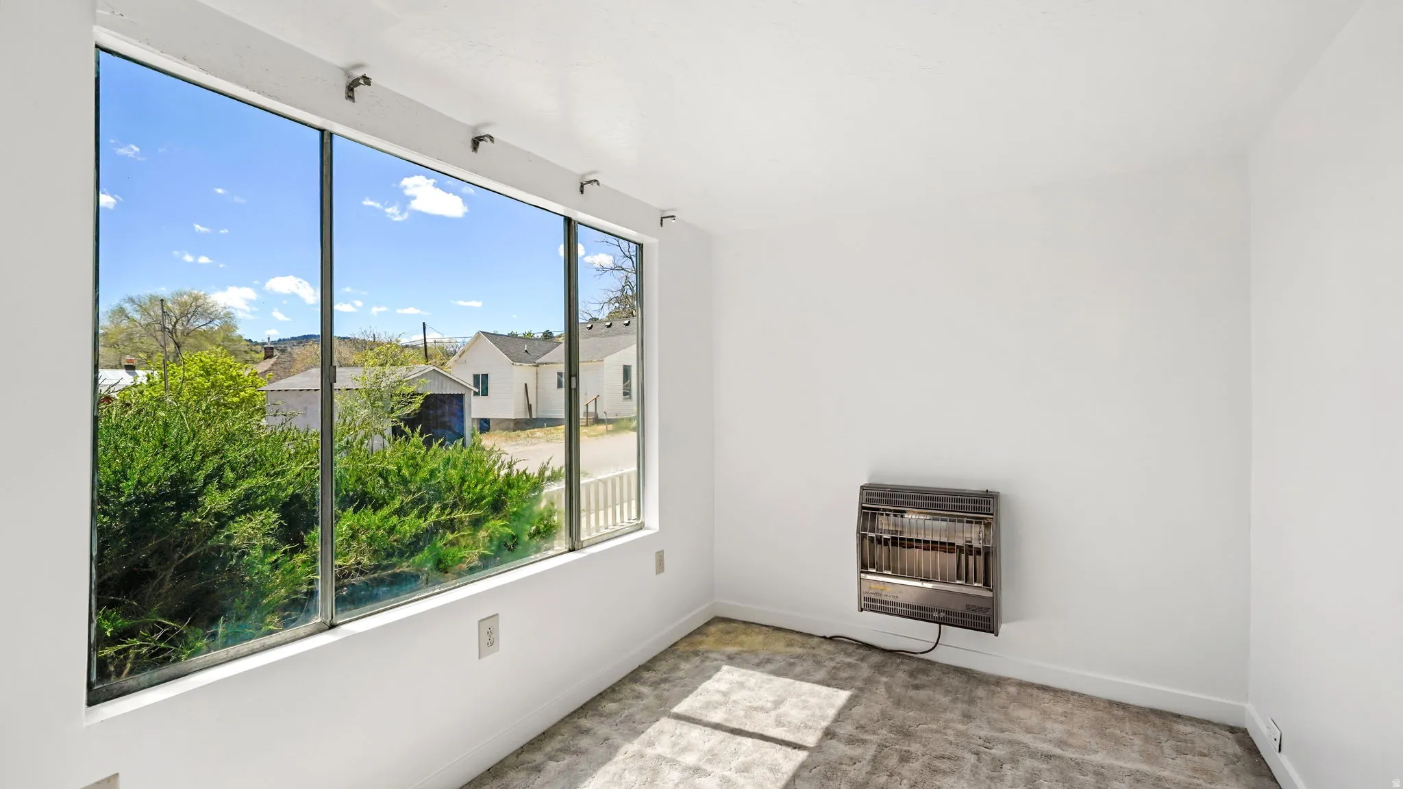 Expansive picture window, white painted walls, wall-mounted heater, light gray textured carpeting, and white baseboards