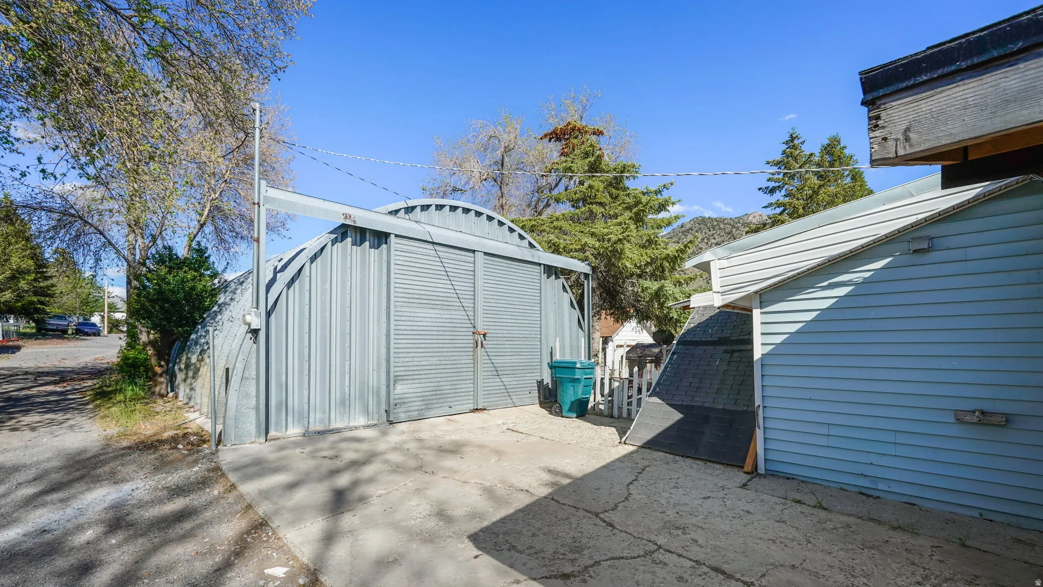 Corrugated metal Quonset hut with roll-up doors, concrete pad, light blue horizontal siding, and an asphalt shingle roof