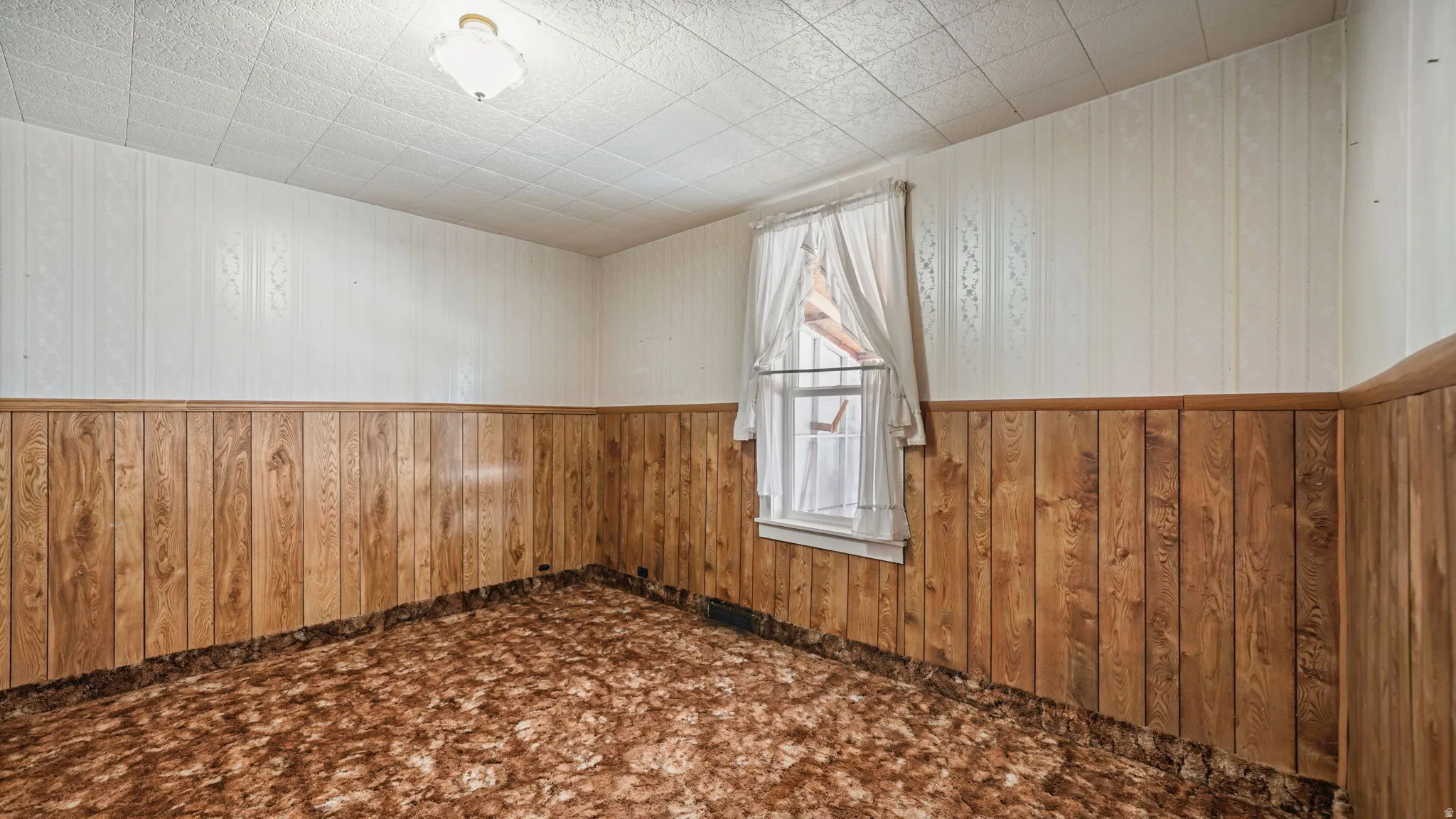 Room featuring wood-finish paneling, patterned wall covering, a grid-pattern ceiling, a single-hung window, and an overhead light fixture
