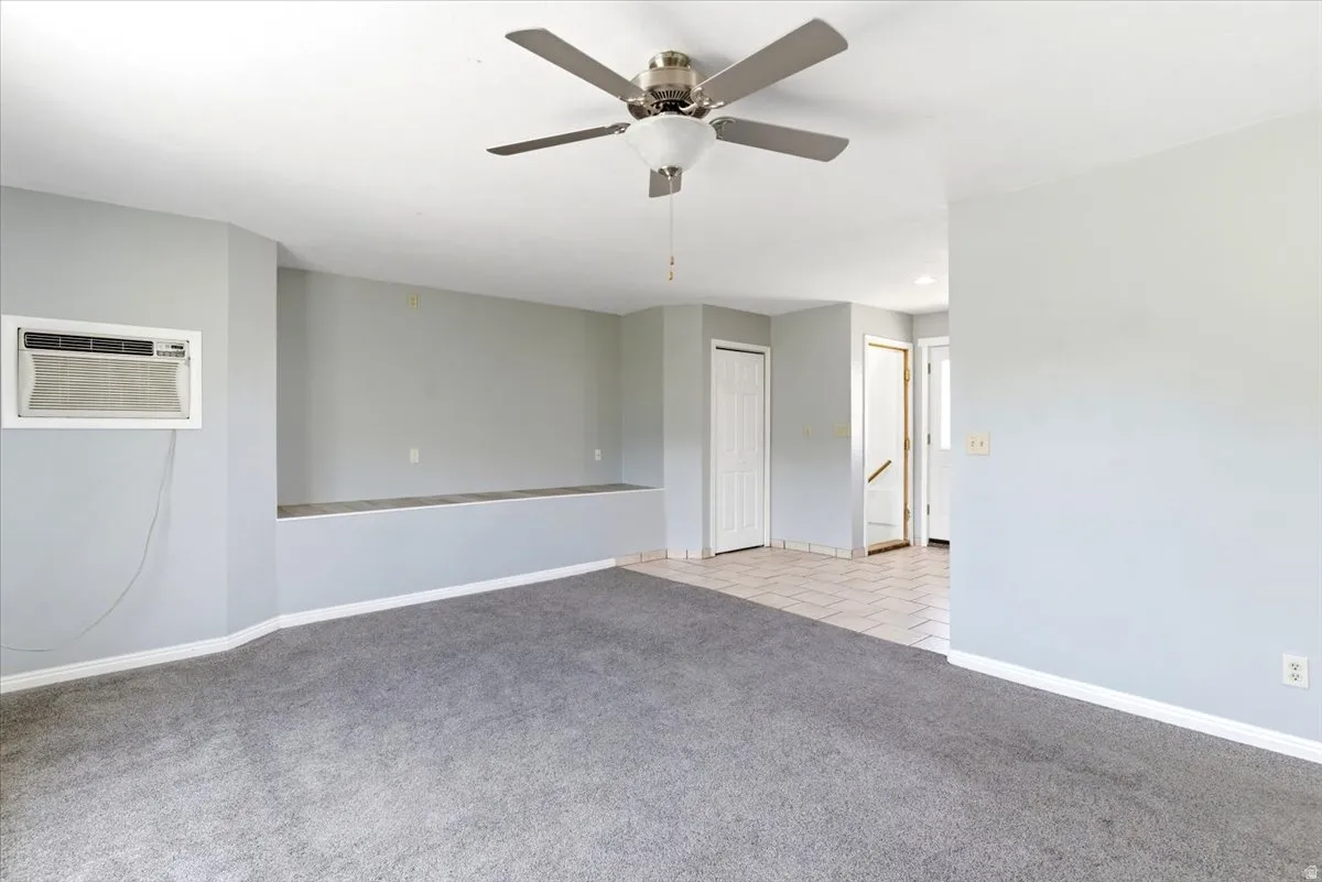 Spacious living area featuring gray carpeting, light gray walls, a built-in window air conditioning unit, and a ceiling fan with integrated lighting