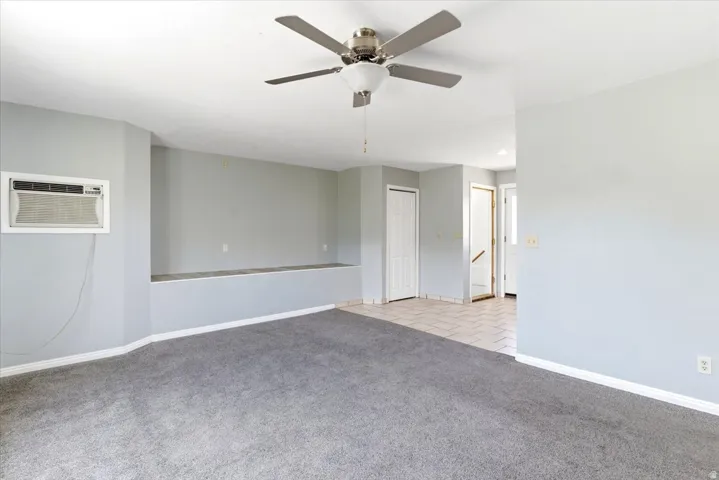 Spacious living area featuring gray carpeting, light gray walls, a built-in window air conditioning unit, and a ceiling fan with integrated lighting
