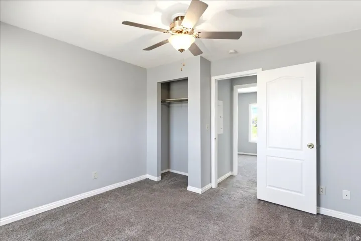 Interior room with light gray walls, gray carpeting, a ceiling fan with light fixture, an open closet with shelving and hanging rod, and a white interior door with brass hardware
