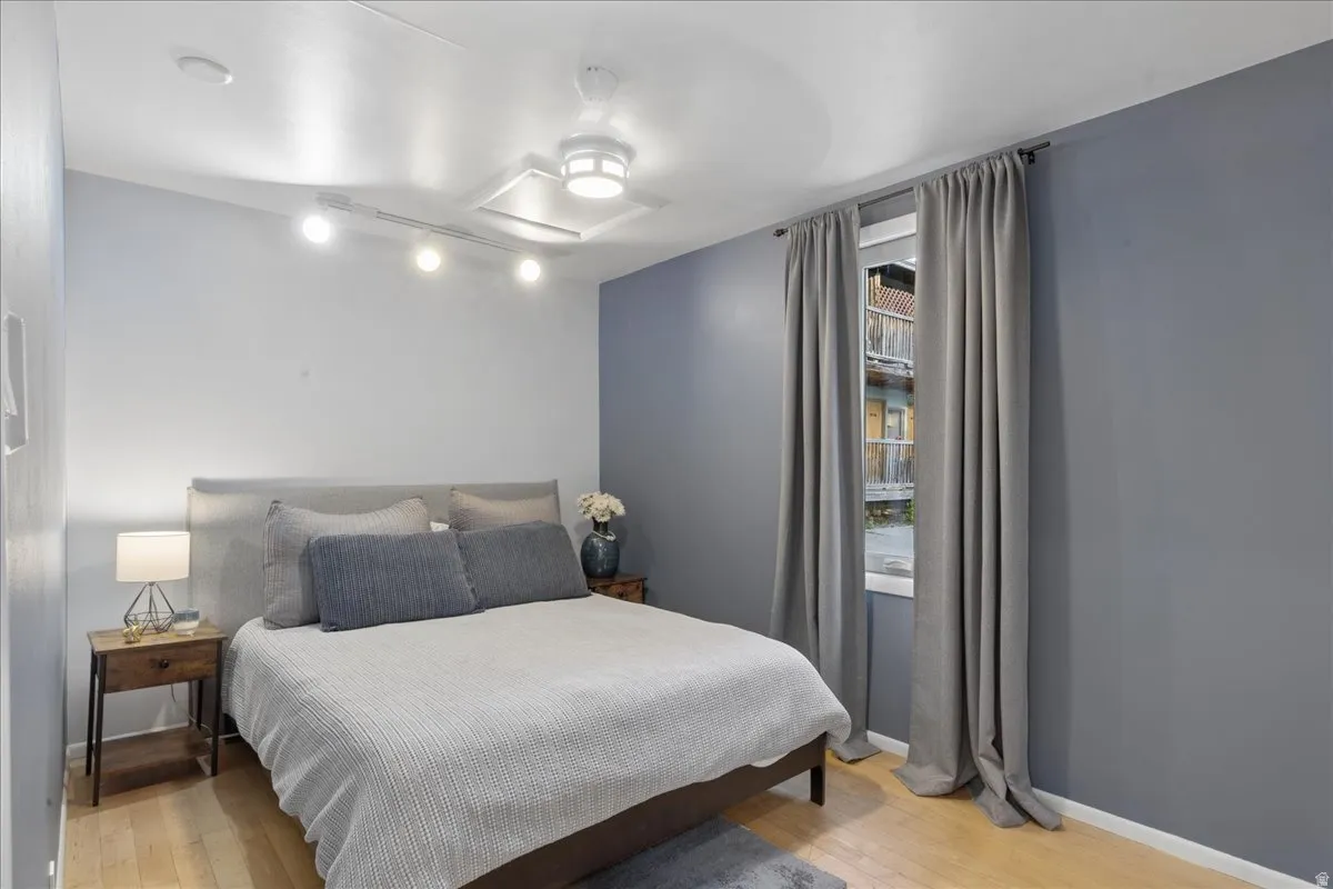 Bedroom featuring light wood-finish flooring, a window with drapery rods, track lighting, and a ceiling fan with integrated lighting