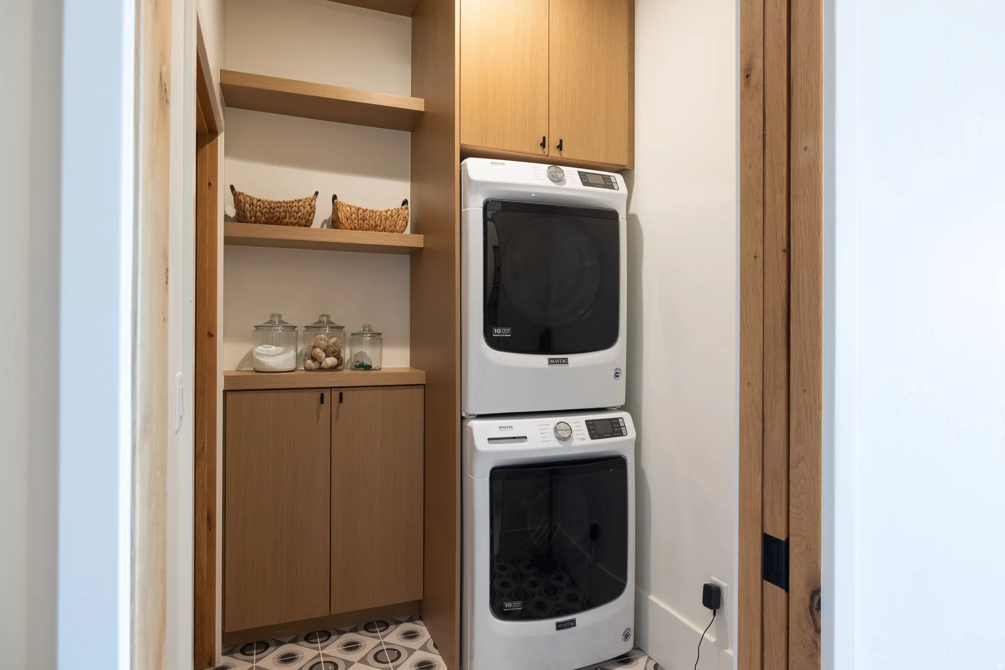 Dedicated laundry area featuring a stacked washer and dryer, wood-finish cabinetry, open shelving, and patterned floor tiling