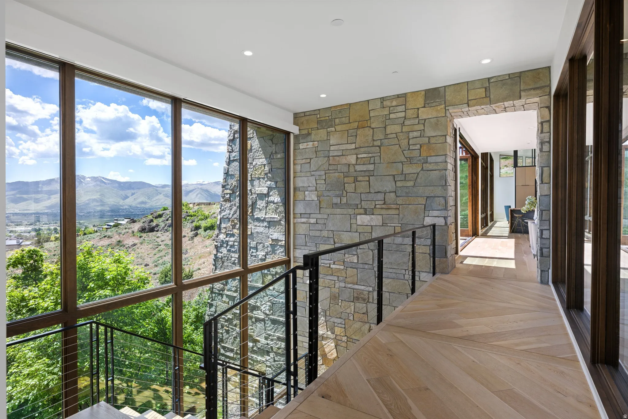 Bright hallway with expansive floor-to-ceiling windows, stone accent wall, chevron wood-finish flooring, recessed lighting, and a contemporary metal and cable railing system