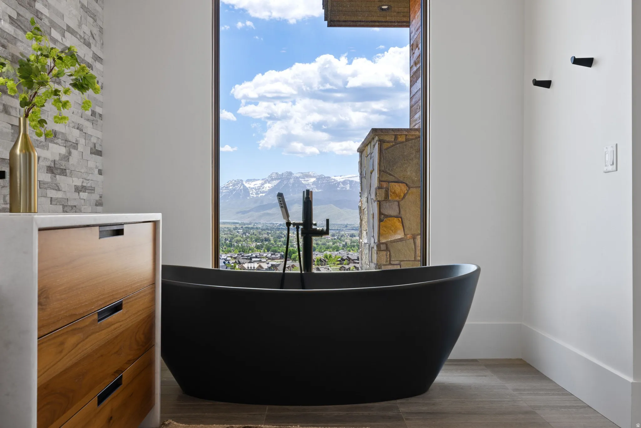 Freestanding black soaking tub with a floor-mounted faucet, framed by a large picture window offering a mountain and city backdrop