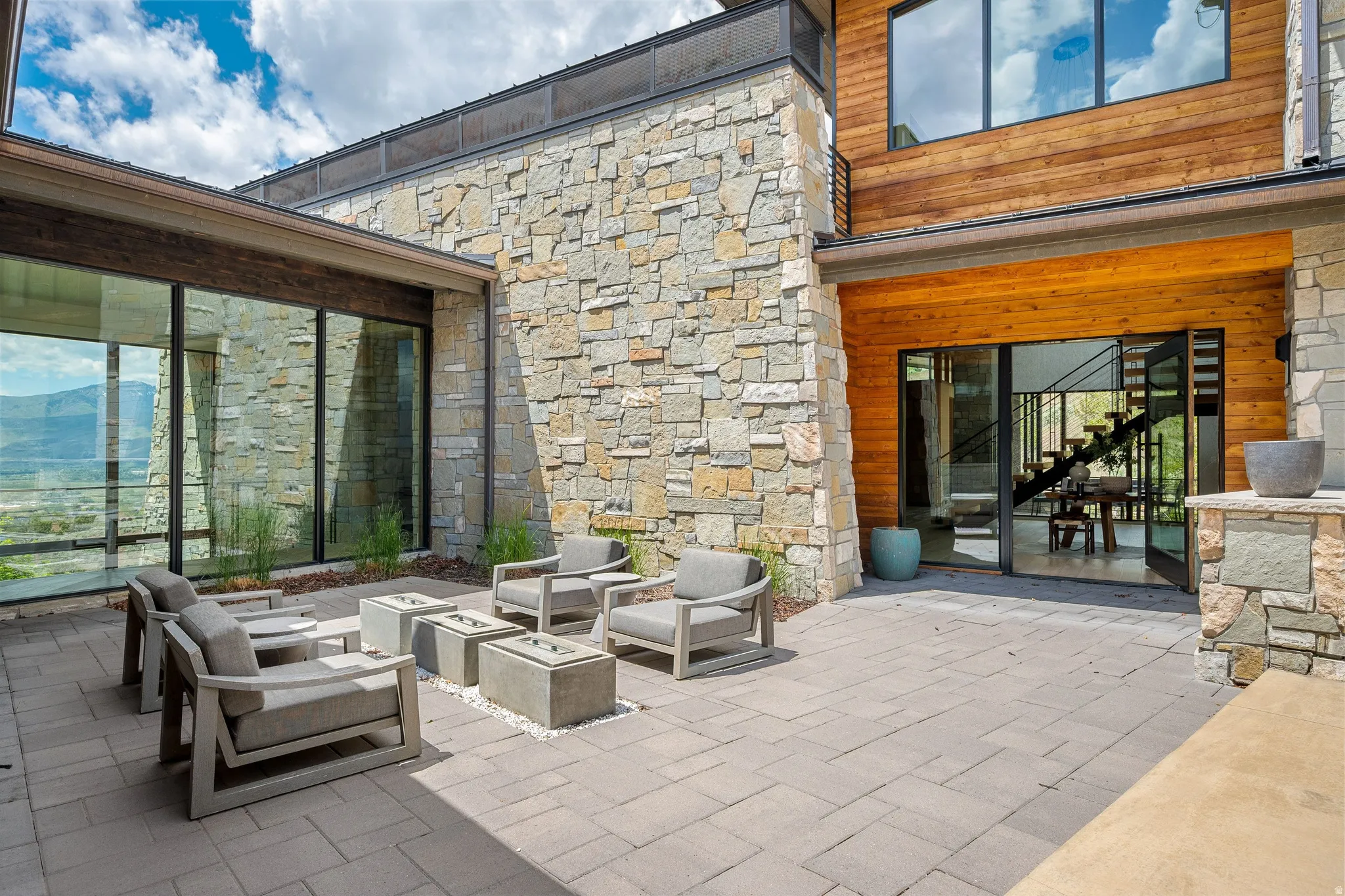 Expansive outdoor patio with large format pavers, stone veneer exterior, wood paneling accents, floor-to-ceiling windows, and multi-panel sliding glass doors