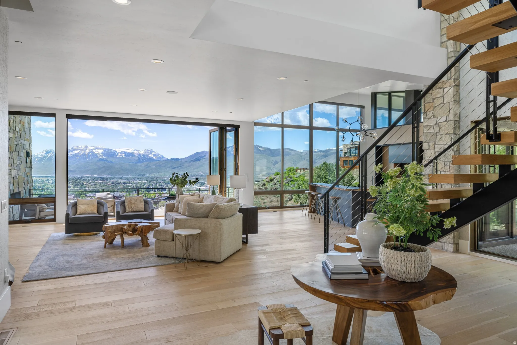 Spacious living area featuring large-format accordion glass doors, light wood-finish flooring, a stone-clad accent wall, open-tread stairs with a metal railing, and abundant natural light