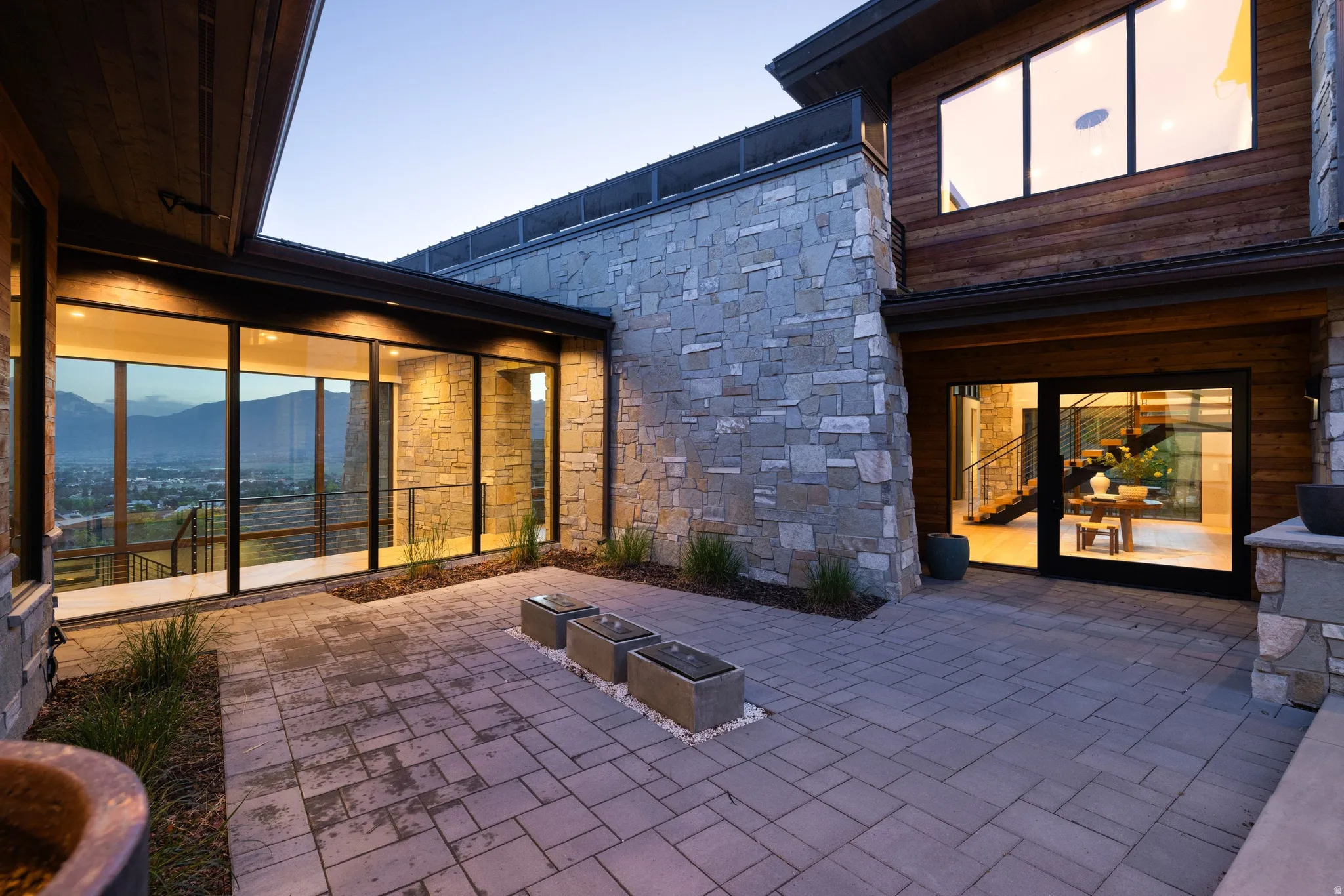 Exterior paver patio featuring stone and wood siding, expansive floor-to-ceiling windows, and integrated outdoor lighting