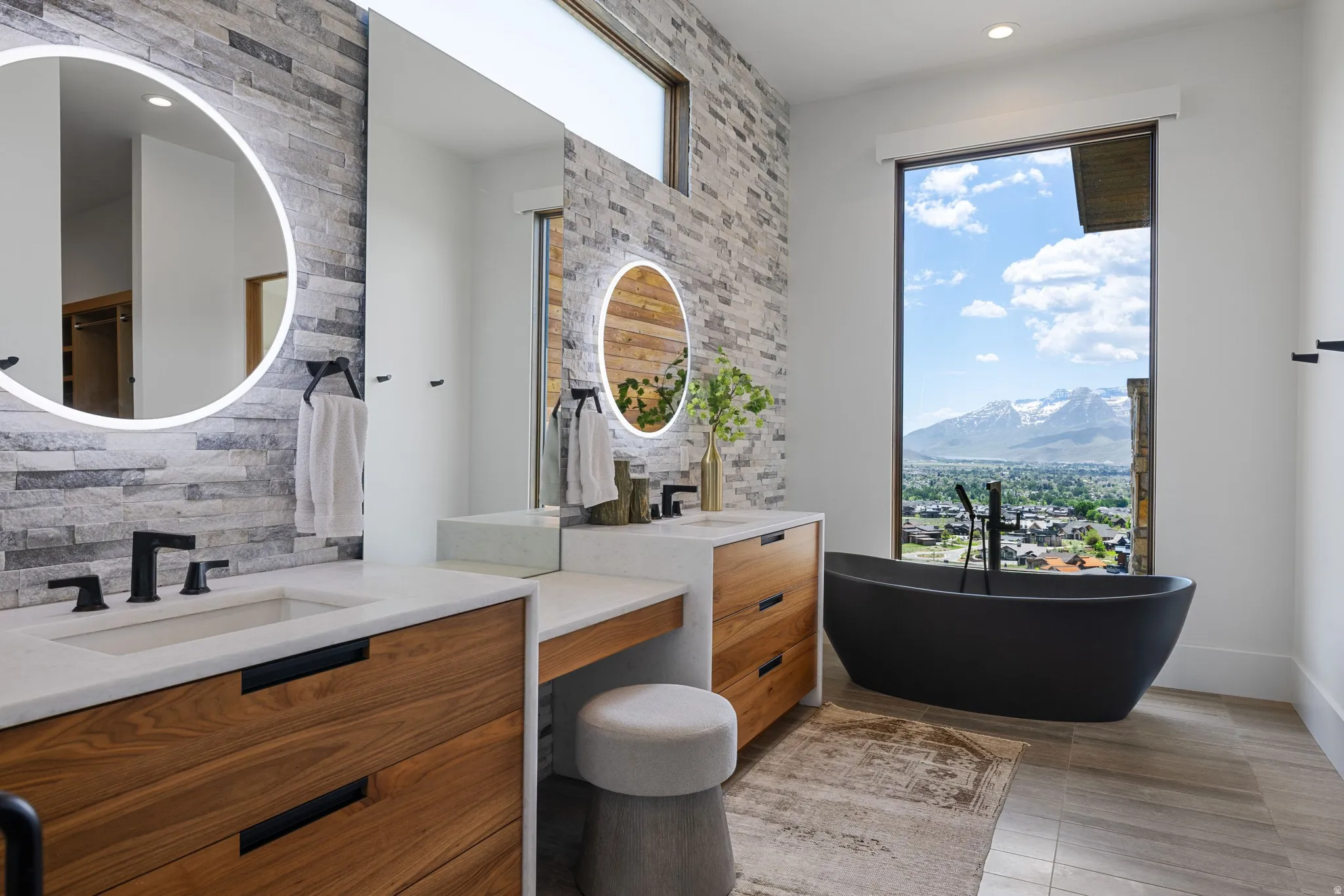 Modern bathroom featuring a black freestanding bathtub with floor-mounted faucet, a vanity with wood-finish cabinetry, integrated sinks, and backlit circular mirrors, and a stone accent wall