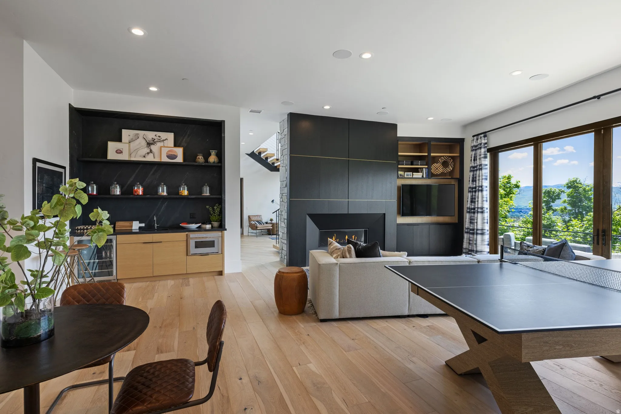 Spacious entertainment area featuring wood-finish flooring, a built-in wet bar with a wine refrigerator and microwave, a modern fireplace with dark paneling, an integrated media center with shelving, and large glass doors opening to a scenic mountain back