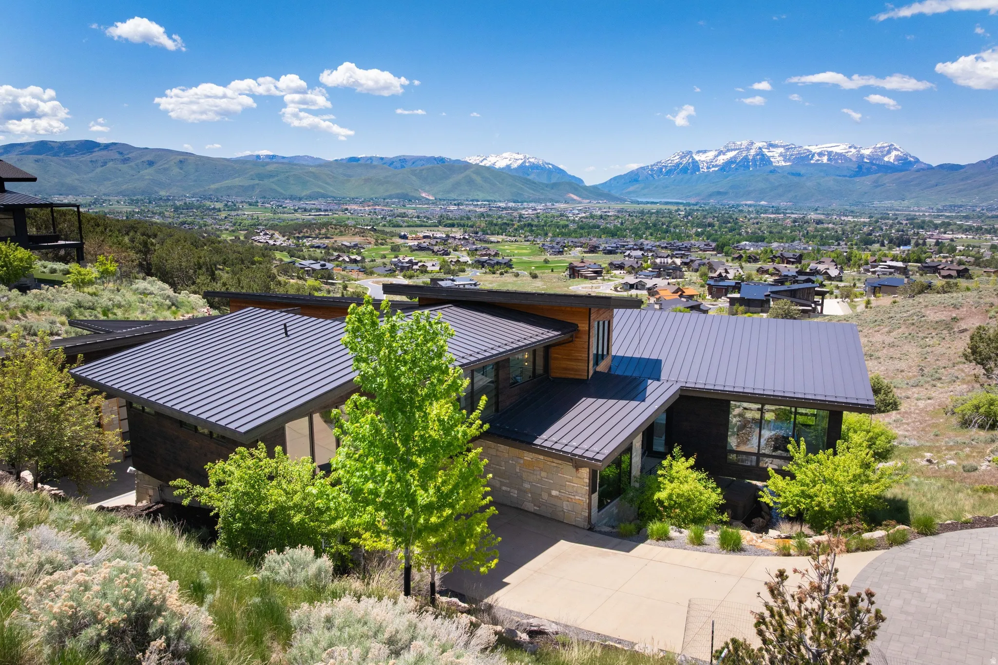 Modern architectural residence featuring multi-pitched metal roofing, dark-toned siding, stone accents, expansive window placements, and a concrete driveway