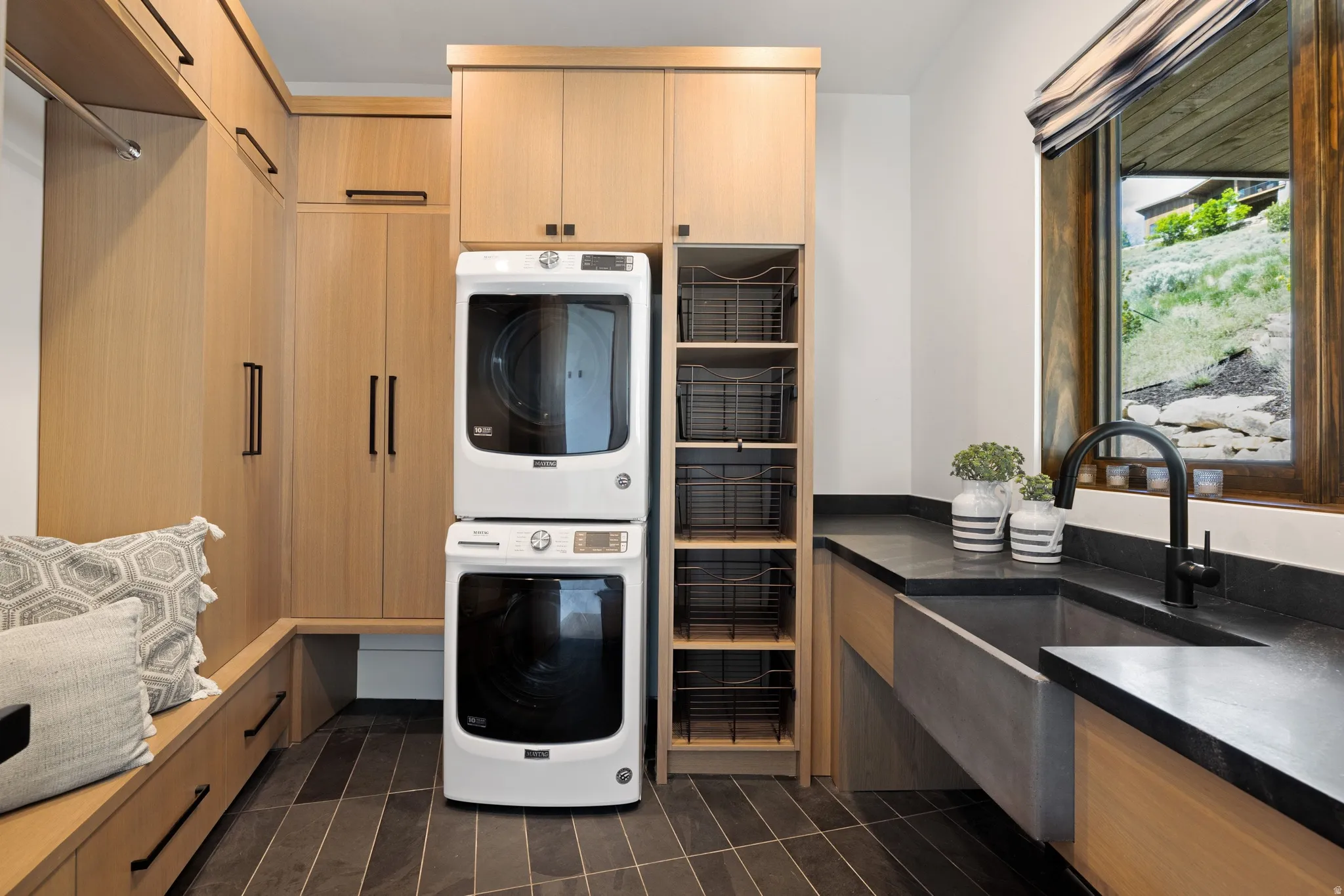 Laundry room featuring extensive built-in light wood cabinetry, integrated washer and dryer, dark stone countertops, a large utility sink with a matte black faucet, and slate-finish floor tiling