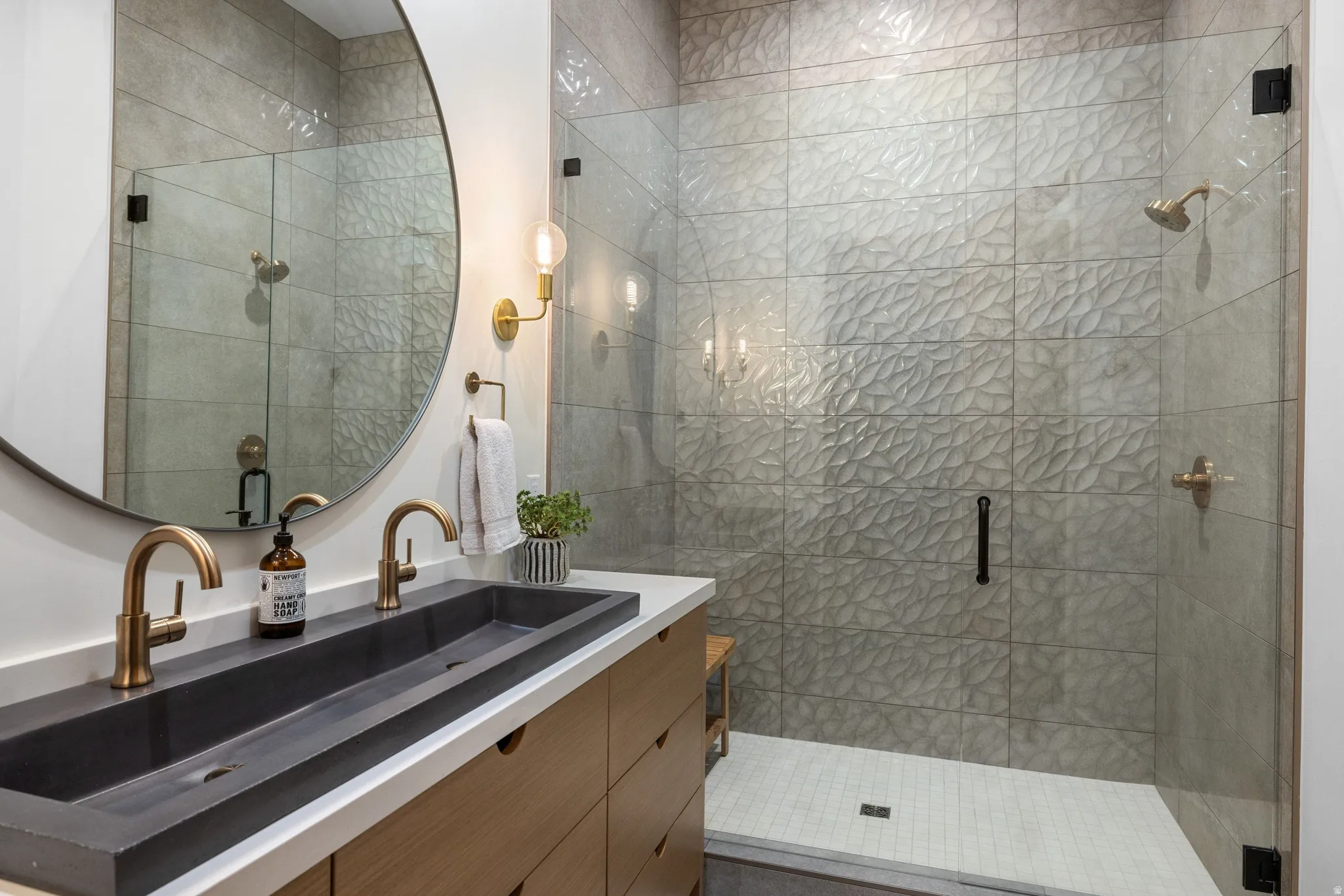 Contemporary bathroom featuring a dual-sink vanity with a dark basin, gold-toned fixtures, a circular mirror, and a glass-enclosed shower with textured tile