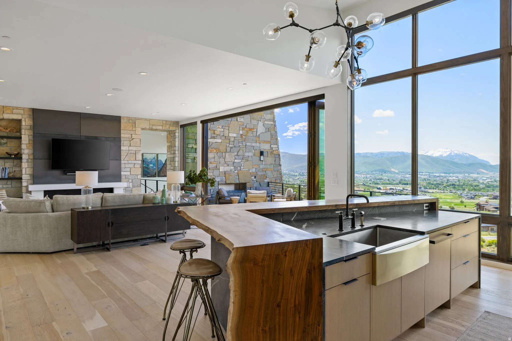 Open-concept living space featuring expansive floor-to-ceiling windows, light wood-finish flooring, a stone accent wall, a built-in media console, and a kitchen island with a natural-edge wood bar top and an integrated farmhouse sink