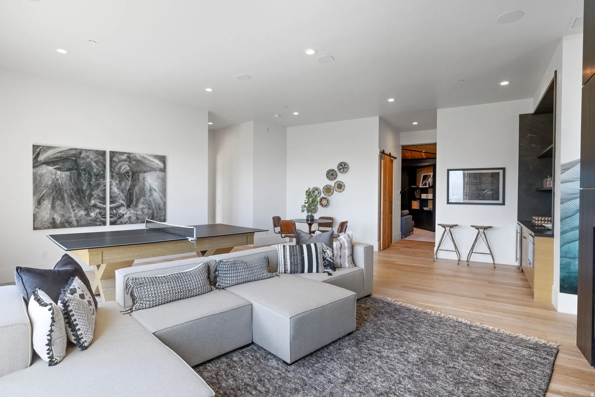 Spacious open-concept room featuring light wood-finish flooring, recessed lighting, a built-in wet bar with dark stone countertop, and a sliding barn door entryway