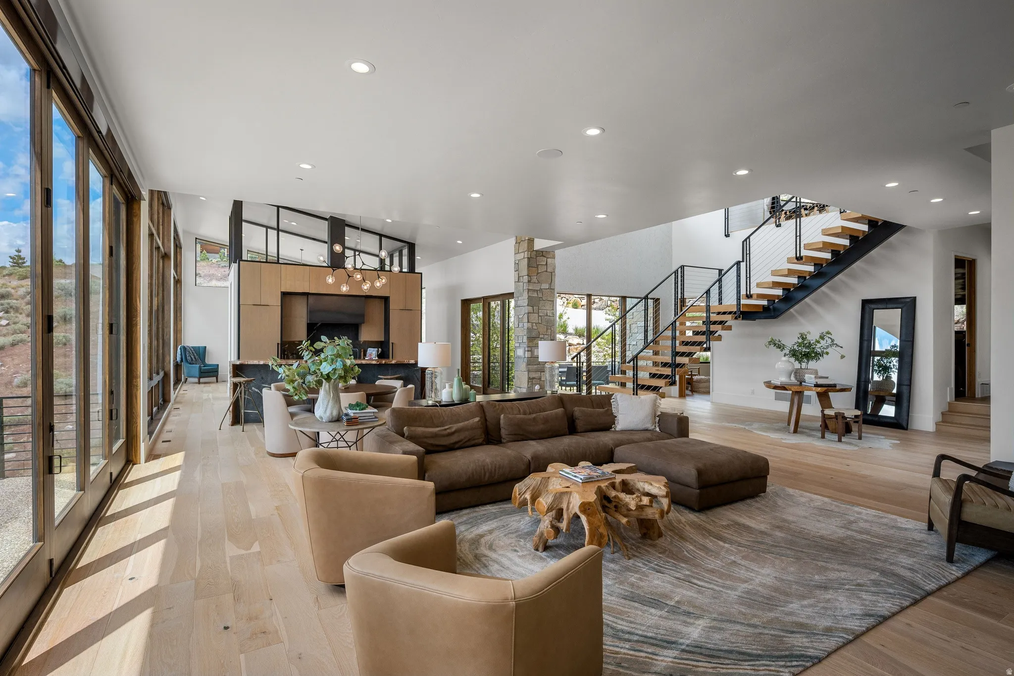 Spacious great room featuring light wood-finish flooring, a contemporary floating staircase with metal railings, a stone-clad pillar, recessed ceiling lighting, and expansive glass doors