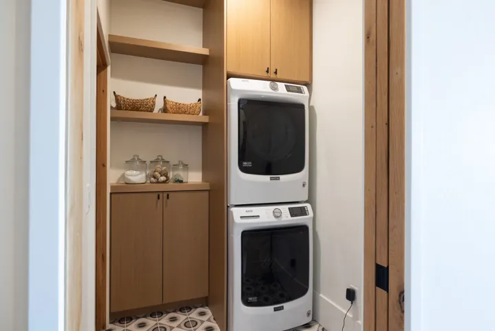 Dedicated laundry area featuring a stacked washer and dryer, wood-finish cabinetry, open shelving, and patterned floor tiling