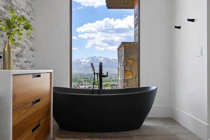 Freestanding black soaking tub with a floor-mounted faucet, framed by a large picture window offering a mountain and city backdrop