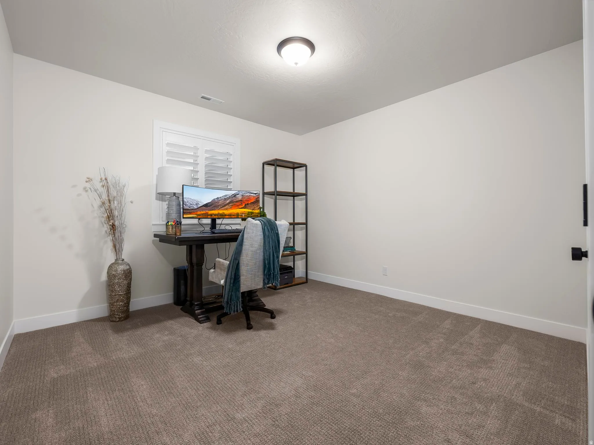 Neutral wall paint complementing plush carpeting, featuring a window with plantation shutters and a ceiling-mounted light fixture