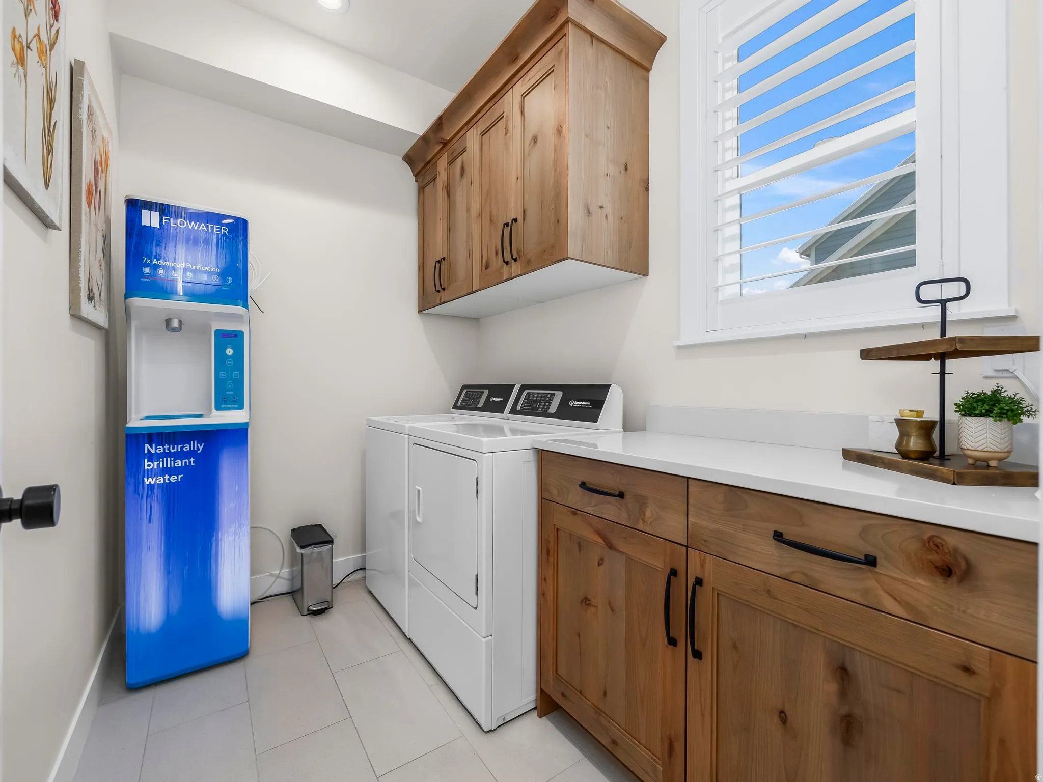Utility space featuring light wood-finish cabinetry, white countertops, a white-framed window with plantation shutters, light tile flooring, and integrated laundry appliances