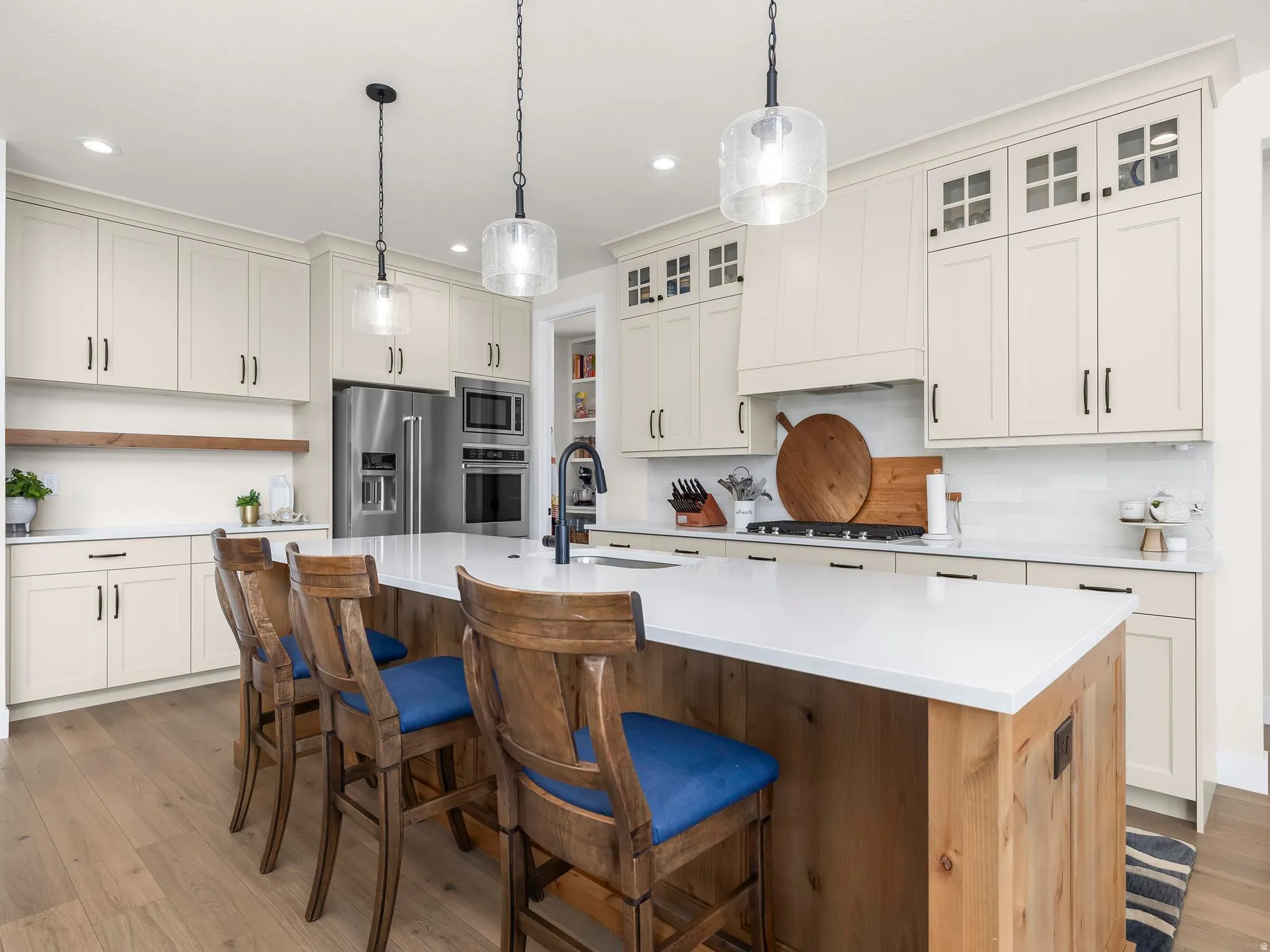 Kitchen featuring an expansive center island with a light wood base and white countertop, stainless steel appliances, light-toned cabinetry with black hardware, a white tile backsplash, and wood-finish flooring