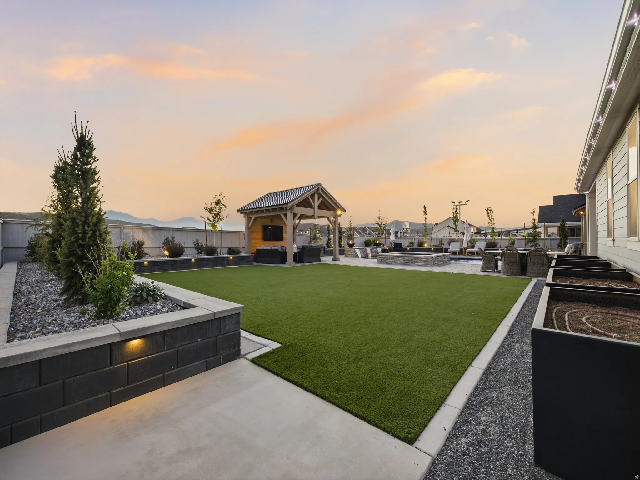 Expansive backyard featuring a lush green lawn, an outdoor kitchen under a wood-beamed pergola, a stone fire pit area, and raised garden beds