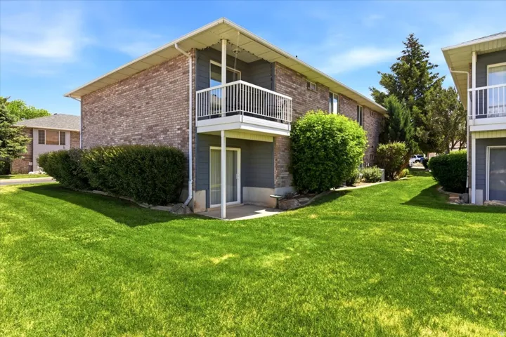Exterior featuring brick siding, a private balcony with white railings, a ground-level patio, mature shrubbery, and a manicured lawn
