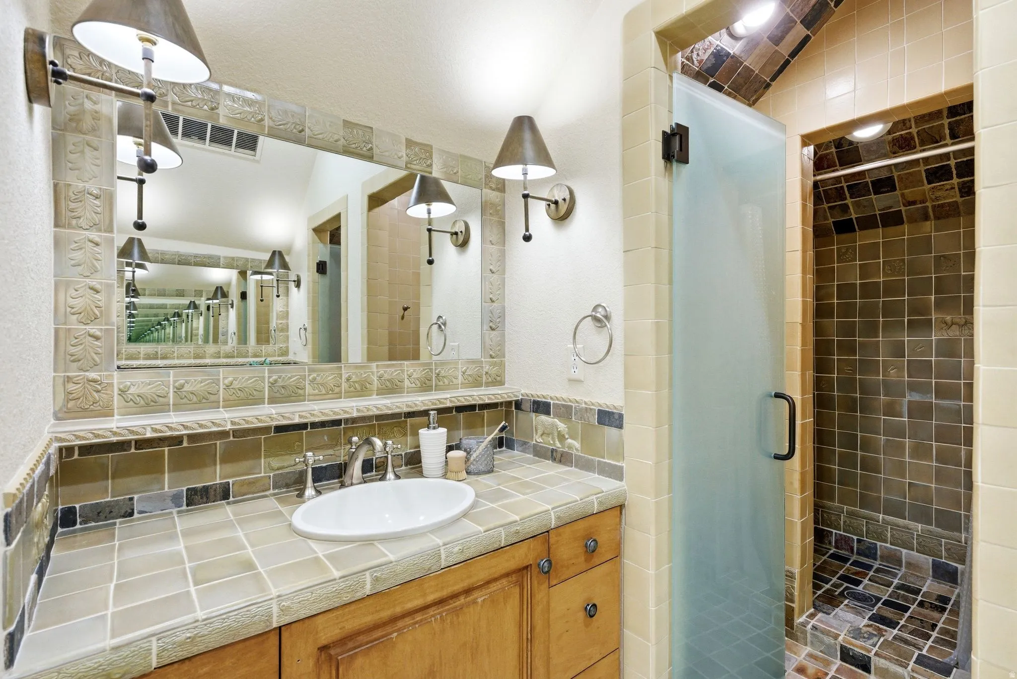 Bathroom featuring a wood-finish vanity with an oval basin, a tile countertop, and a full-width mirror with sconce lighting