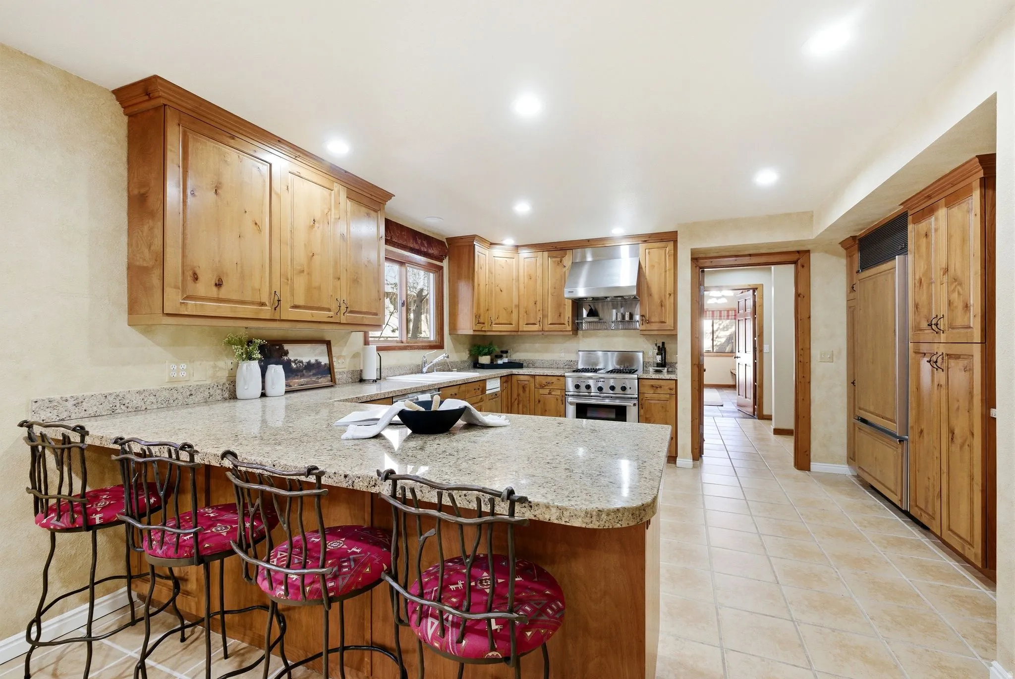 Kitchen featuring a large central island with a light-toned countertop, extensive wood cabinetry, stainless steel range with overhead vent, integrated refrigerator, and light tile flooring