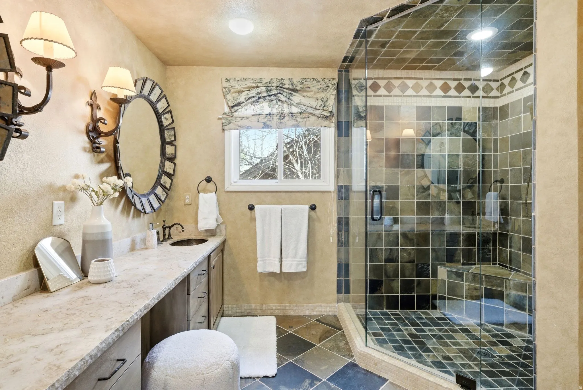 Bathroom featuring a glass-enclosed shower with tile surround and integrated bench seating, a dual-sink vanity with stone countertop, and an oval mirror