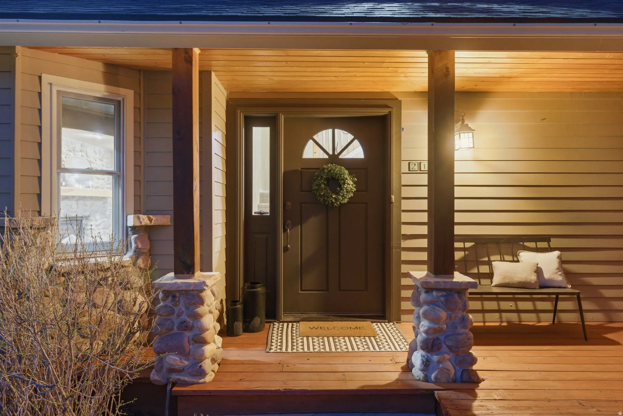 Inviting front porch featuring a dark entry door with a transom window, a sidelite, wood-finish flooring, and natural stone-clad columns