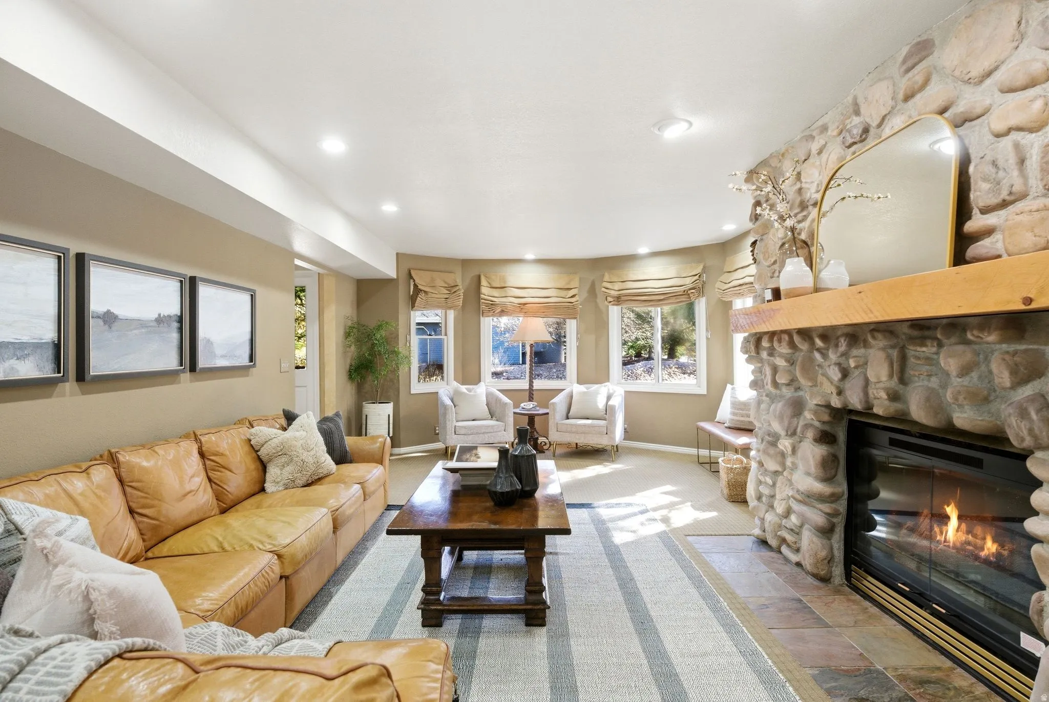 Spacious living area featuring a prominent stone fireplace with a wood mantle, recessed lighting, and multiple windows with Roman shades