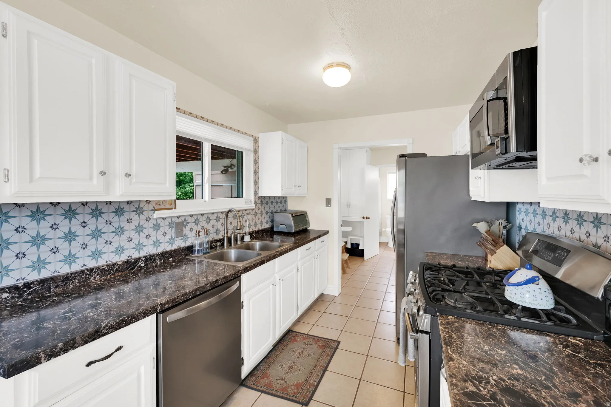 Galley kitchen featuring white cabinetry, patterned tile backsplash, dark countertops, and stainless steel appliances