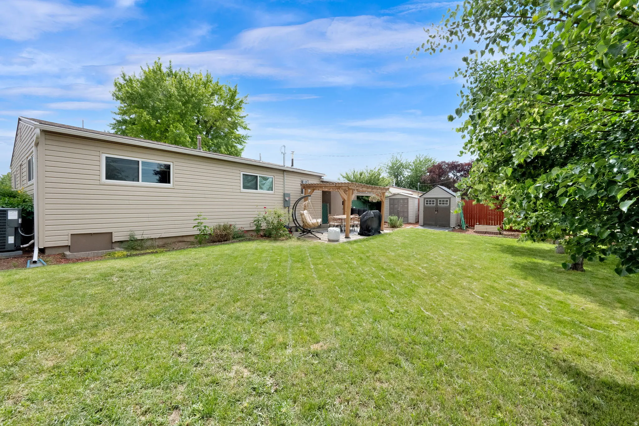 Expansive green lawn with a pergola-covered patio, featuring a wood-sided house, two storage sheds, and mature perimeter trees