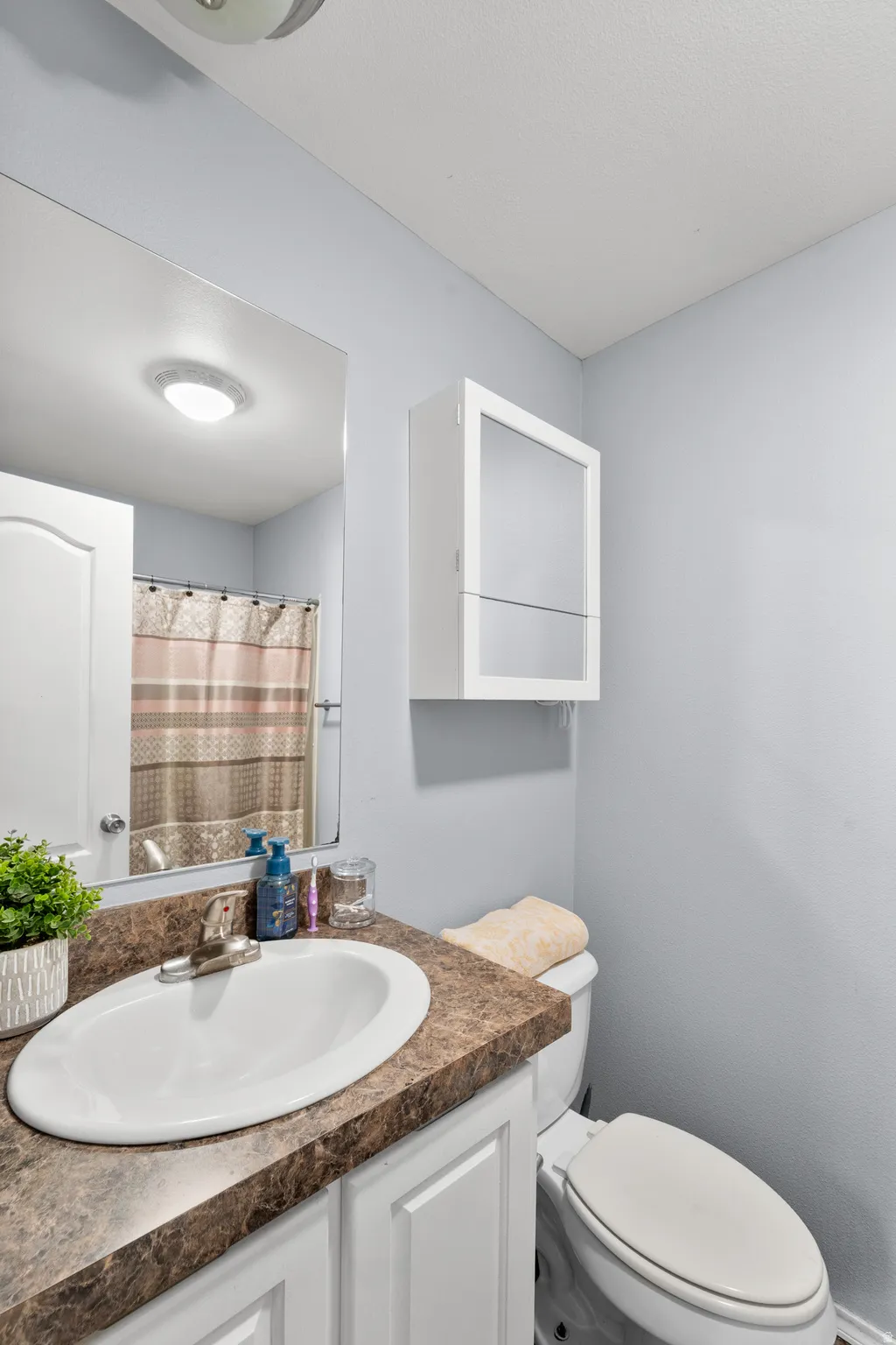Bathroom featuring a single white porcelain sink, a brown patterned countertop, a white vanity with paneling, a wall-mounted mirror, and a white medicine cabinet