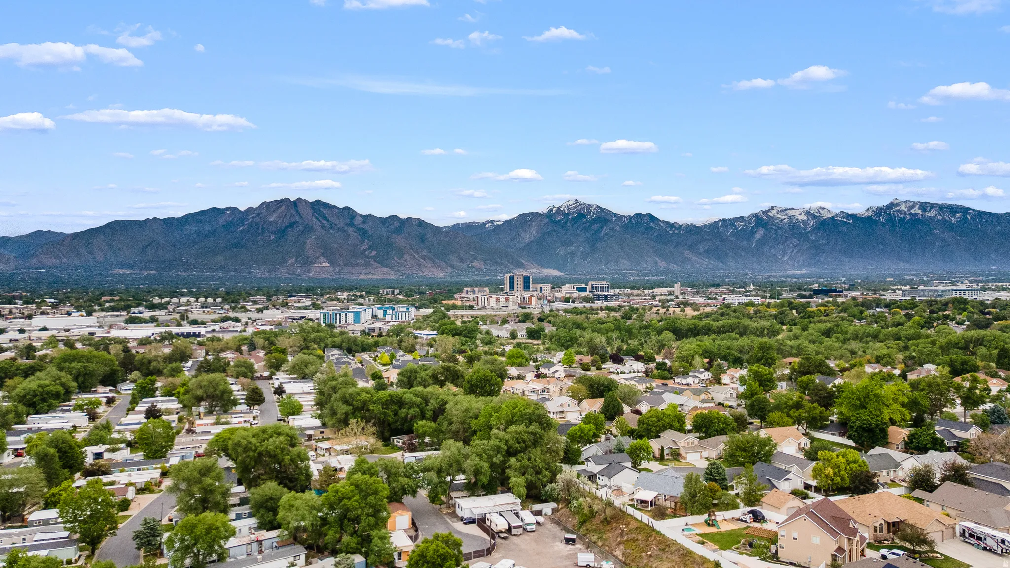 Expansive aerial perspective showcasing a residential area with mature trees, a distant mountain range with snow-capped peaks, and a cluster of urban buildings on the horizon