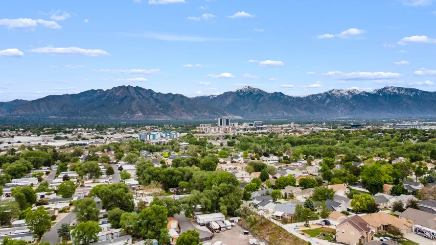 Expansive aerial perspective showcasing a residential area with mature trees, a distant mountain range with snow-capped peaks, and a cluster of urban buildings on the horizon