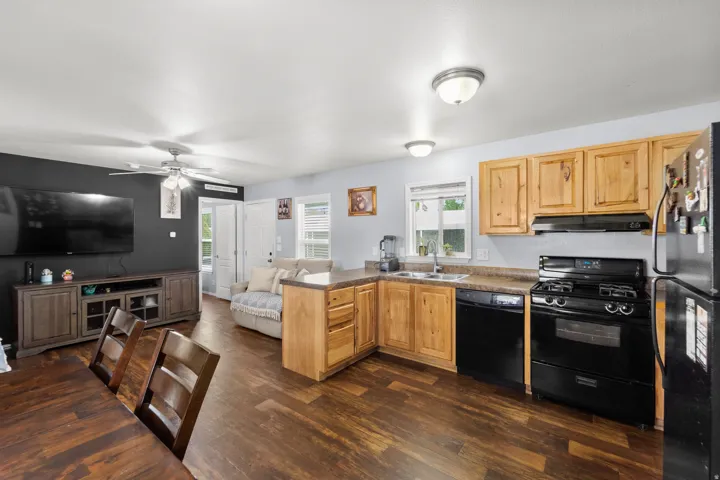 Open-concept living area featuring wood-finish flooring throughout, a kitchen with natural wood cabinetry and black appliances, a ceiling fan with integrated lighting, and recessed ceiling lights