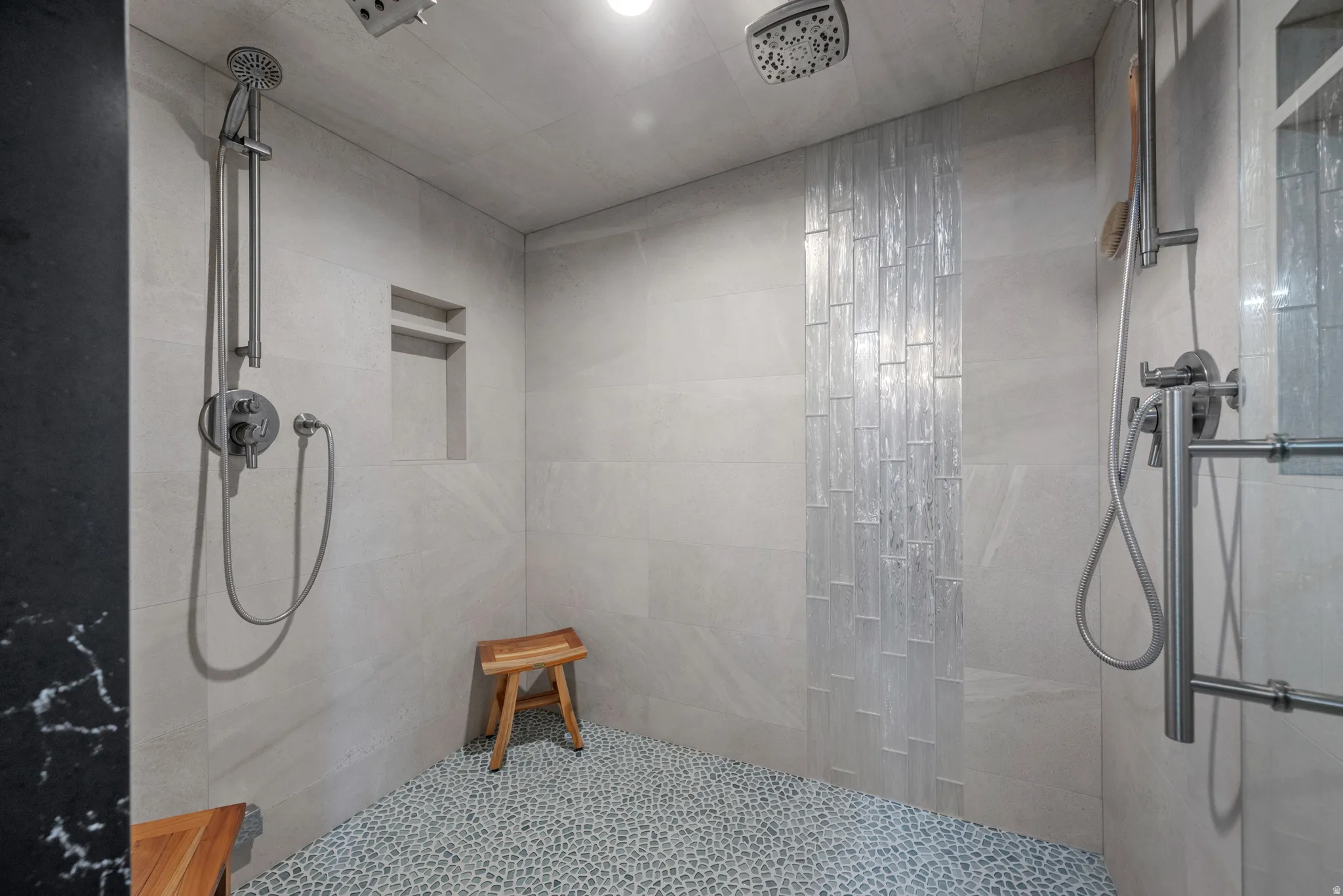Spacious shower featuring a pebble tile floor, light-toned wall tiling, dual showerheads, and an integrated wall niche