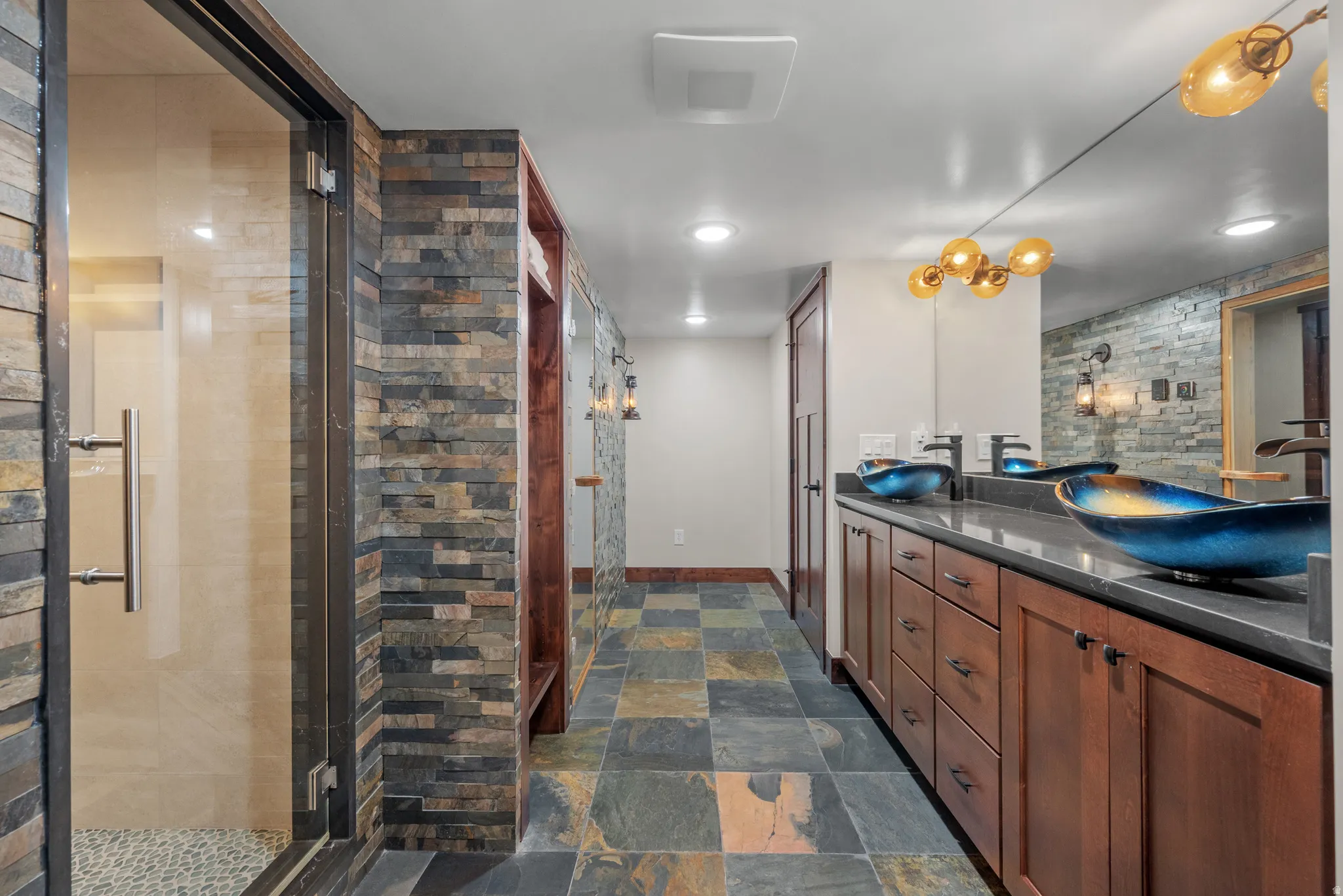 Double vanity with vessel sinks, slate tile flooring, walk-in shower with glass enclosure, stacked stone accent walls, and custom wood cabinetry