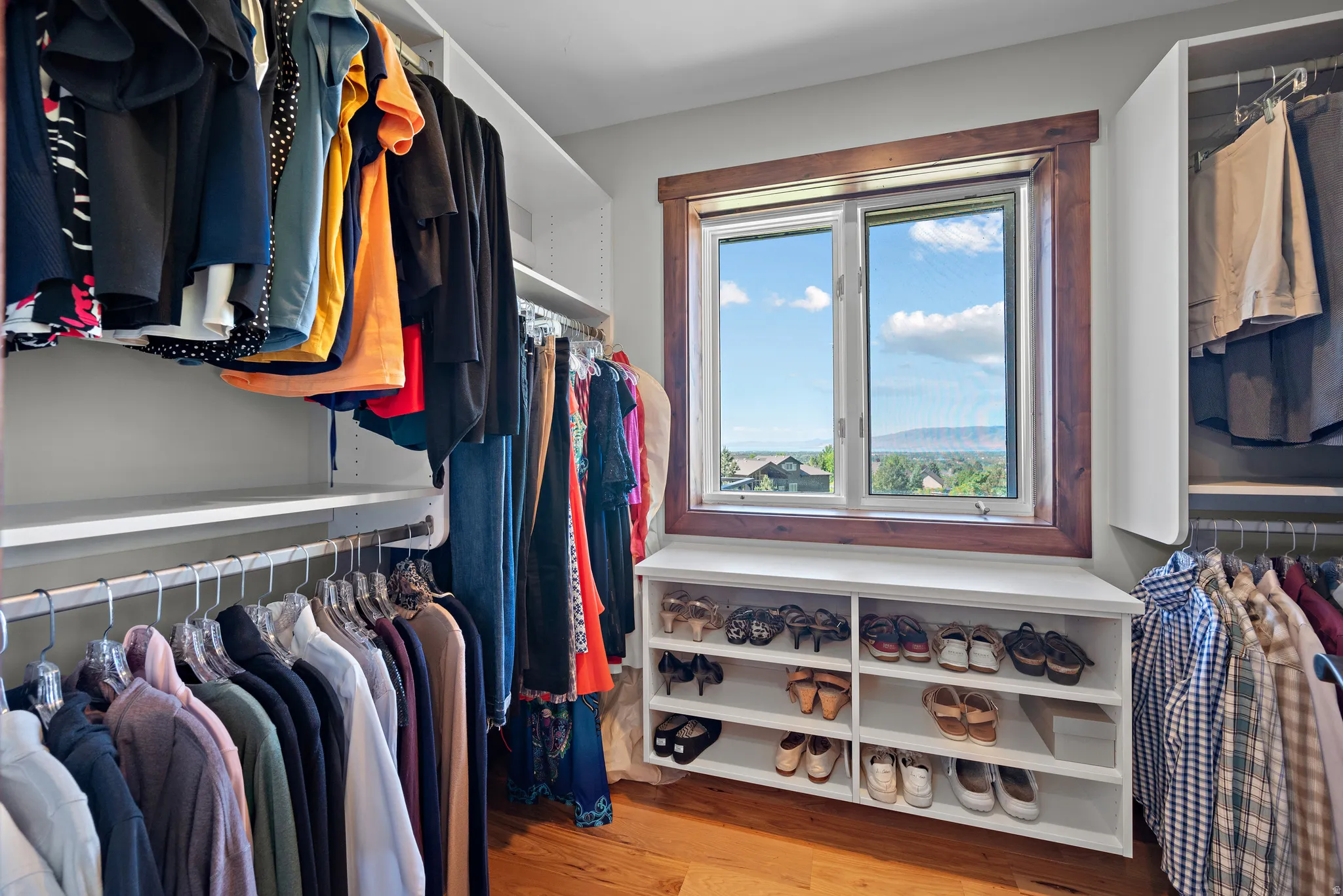 Custom walk-in closet featuring extensive built-in shelving and hanging rods, a built-in window seat with shoe storage, wood-finish flooring, and a large window with wood trim